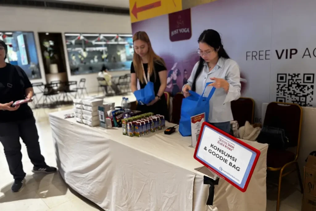 Meja konsumsi dan goodie bag dalam acara Donor Darah Rumah Berkat ke-9, dengan staf yang sedang mempersiapkan paket untuk peserta.